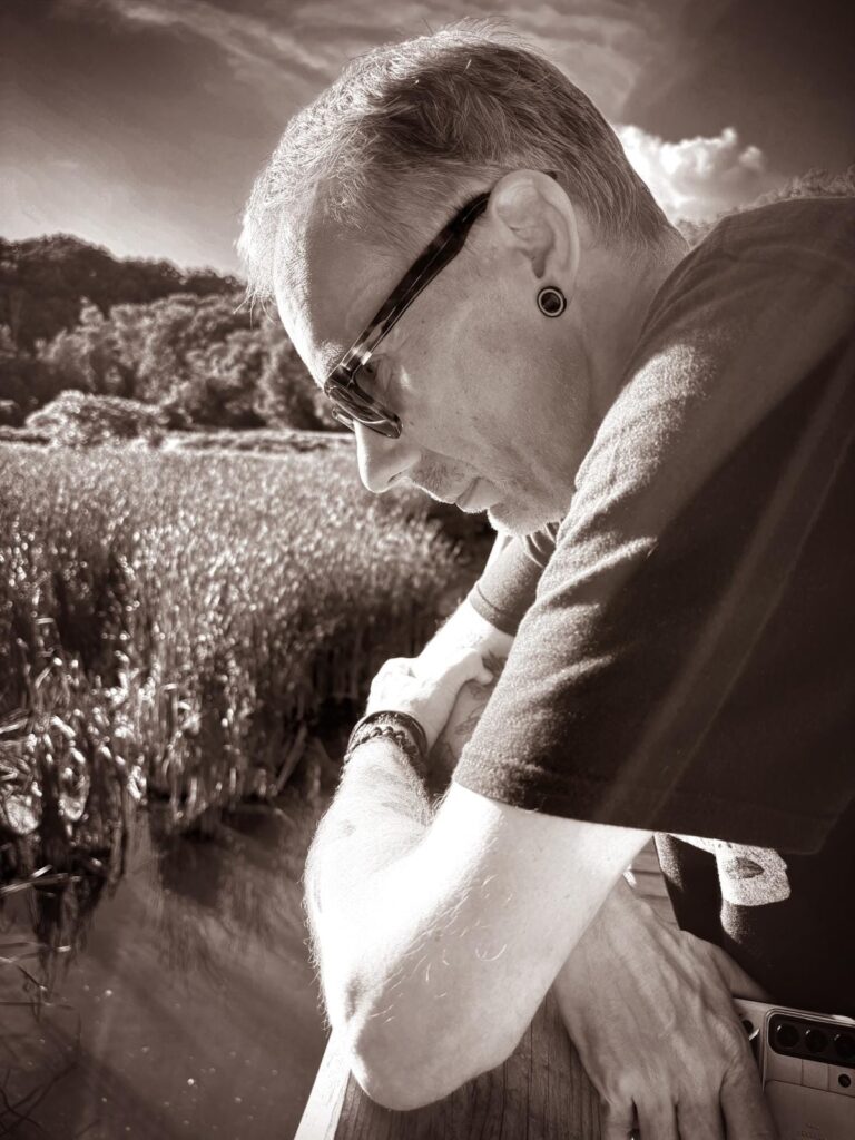 Andygibbphoto sepia image of Andy Gibb in tee shirt and dark glasses leaning over a bridge with arms crossed.