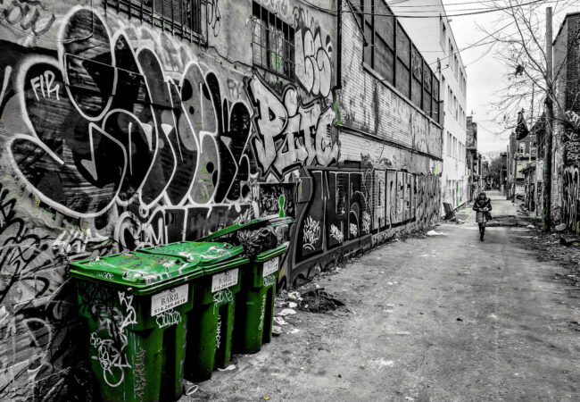 An Andygibbphoto black and white image of lane in Montreal with walls covered in graffiti and wall art, there are also three bins in the fore ground coloured green in the image, and a woman walking down the lane